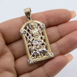 May include: A gold and silver pendant featuring a detailed depiction of Saint Lazarus, surrounded by a decorative, intricate lattice design. The pendant has a bail for attaching to a chain.