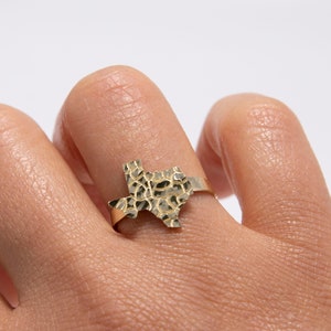 May include: A gold ring shaped like the state of Texas. The ring has a textured surface on the Texas shape. The ring is displayed on a finger. The background is white.