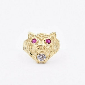 Op de afbeelding: Een gouden ring met een tijgerkopontwerp. De tijger heeft rode ogen en een witte steen in het midden van zijn kop.