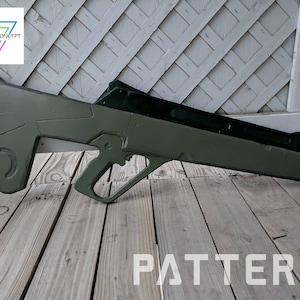 May include: A green, futuristic-looking rifle prop with a black handle and a textured surface. The prop is lying on a wooden surface. The word "PATTERN" is printed in white on the surface.