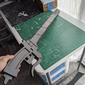 May include: A gray foam sword with a detailed design, resting on a green cutting mat. The sword is partially assembled and has a handle and a blade.