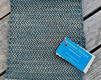 May include: A square, handwoven textile in a blue and cream herringbone pattern. The fabric has a textured appearance. A blue tag with the text "CIMARRON TEXTILES Handmade by Kim" is attached.