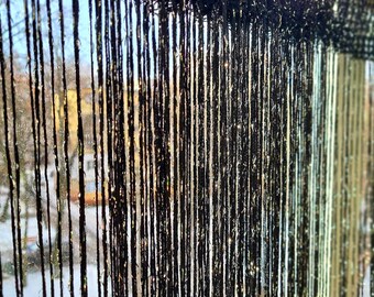 May include: Black, vertical string curtain hanging in front of a window. The strings are closely spaced, creating a textured effect. The window shows a view of a snowy landscape and buildings.