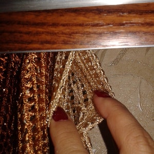 May include: Close-up of a gold and brown crocheted curtain with a hand reaching out to touch it.
