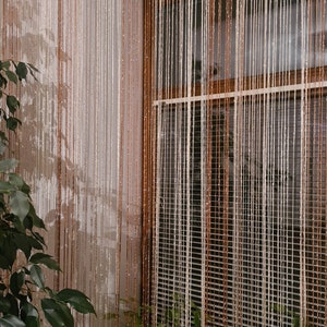 May include: A window with a white and brown beaded curtain. The curtain is made of thin strings of beads that hang from a rod. There are plants in pots on the windowsill.