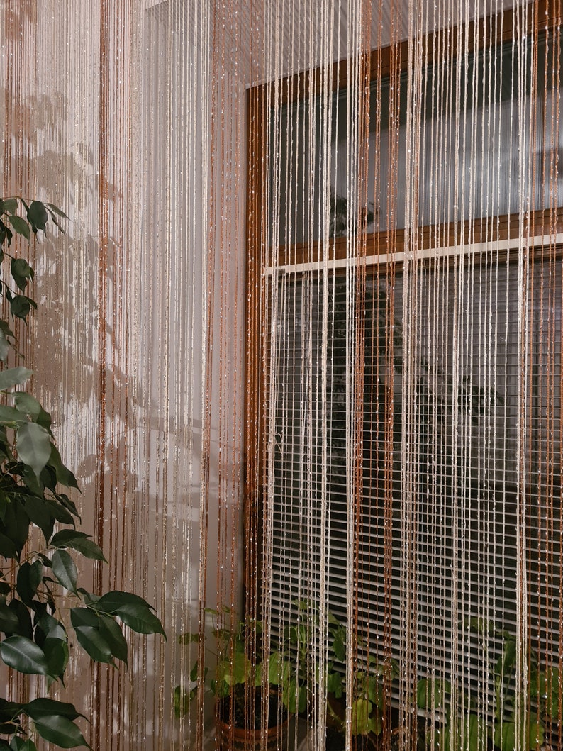 May include: A window with a white and brown beaded curtain. The curtain is made of thin strings of beads that hang from a rod. There are plants in pots on the windowsill.