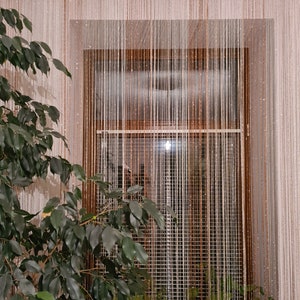 May include: A window with a sheer beaded curtain in white and brown. The curtain is made of thin strings and hangs from a wooden rod. There are plants in pots on the windowsill.