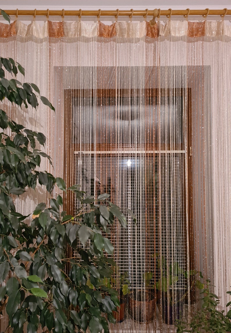 May include: A window with a sheer beaded curtain in white and brown. The curtain is made of thin strings and hangs from a wooden rod. There are plants in pots on the windowsill.