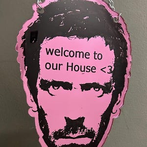 May include: A pink and black acrylic wall hanging shaped like a human head with a beard. The text "welcome to our House <3" is printed in black on the forehead. The piece is suspended by a silver-colored metal chain attached to two top holes.