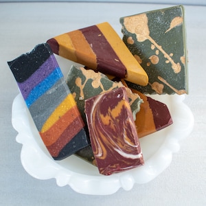 May include: Assortment of six handmade soap bars in a white ceramic dish. The soaps are in various colours and patterns, including purple, blue, yellow, orange, green, brown, and white.