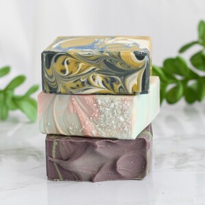 May include: Three bars of handmade soap stacked on top of each other. The top bar is a swirl of black, white, and yellow. The middle bar is a swirl of pink, white, and blue. The bottom bar is a dark purple with a textured surface.
