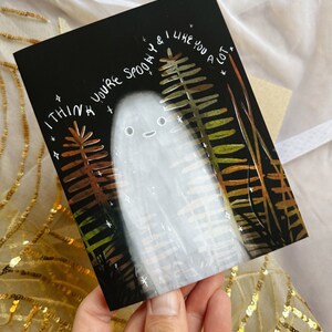 May include: A black card with a white ghost and green, brown, and yellow fern-like plants. The text on the card reads "I think you're spooky & I like you a lot."