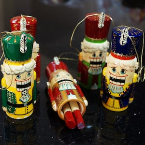 May include: Four wooden nutcracker ornaments with painted details. The nutcrackers are dressed in different colors, including red, green, blue, and yellow. Each nutcracker has a white feather on its hat.