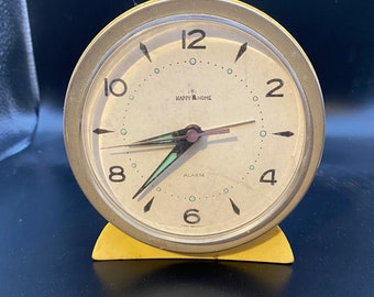May include: A yellow vintage alarm clock with a white face and black hands. The clock face has the words "Happy Home" and "Alarm" printed on it. The clock is made of metal and has a plastic base.