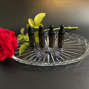 Puede incluir: Cuatro pequeños frascos cuentagotas de vidrio ámbar con tapas negras están colocados en una bandeja de vidrio transparente de forma ovalada. Una rosa roja vibrante y hojas verdes están en la bandeja.