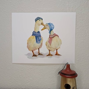 K&ouml;nnte beinhalten: Ein Aquarellbild von zwei Enten, die blaue H&uuml;te und Schals tragen. Die Enten schauen sich an und scheinen sich zu k&uuml;ssen.