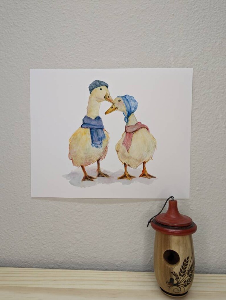 K&ouml;nnte beinhalten: Ein Aquarellbild von zwei Enten, die blaue H&uuml;te und Schals tragen. Die Enten schauen sich an und scheinen sich zu k&uuml;ssen.