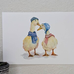 K&ouml;nnte beinhalten: Zwei gelbe Enten, die blaue und rosa Schals und H&uuml;te tragen. Die Enten schauen sich an und scheinen sich zu kuscheln.