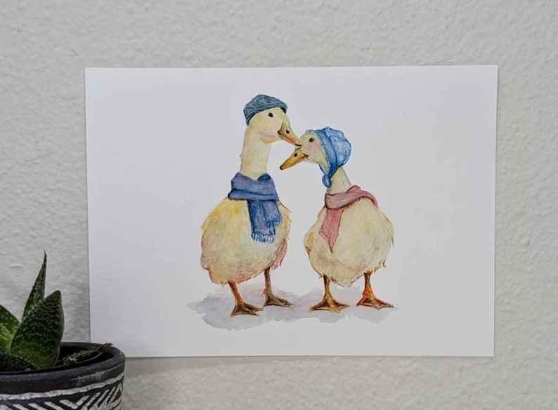 K&ouml;nnte beinhalten: Zwei gelbe Enten, die blaue und rosa Schals und H&uuml;te tragen. Die Enten schauen sich an und scheinen sich zu kuscheln.