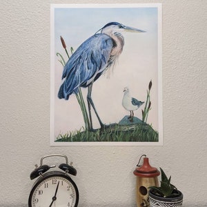 May include: A watercolor painting of a blue heron standing on a rock with a smaller white bird beside it. The heron has a long neck and a long beak. The background is a light blue sky with some green reeds.