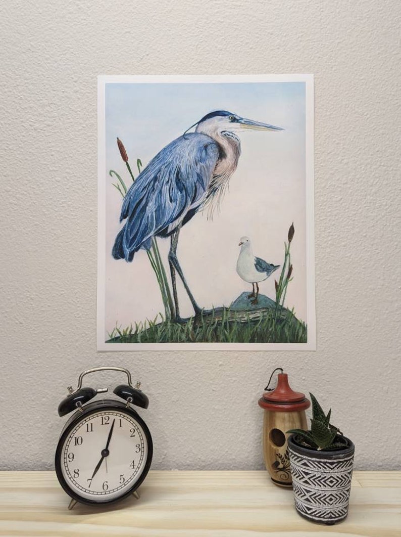 May include: A watercolor painting of a blue heron standing on a rock with a smaller white bird beside it. The heron has a long neck and a long beak. The background is a light blue sky with some green reeds.