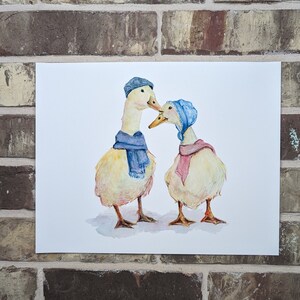 K&ouml;nnte beinhalten: Zwei gelbe Enten, die blaue und rosafarbene Schals und H&uuml;te tragen. Die Enten schauen sich an und scheinen sich zu k&uuml;ssen. Der Hintergrund ist wei&szlig;.