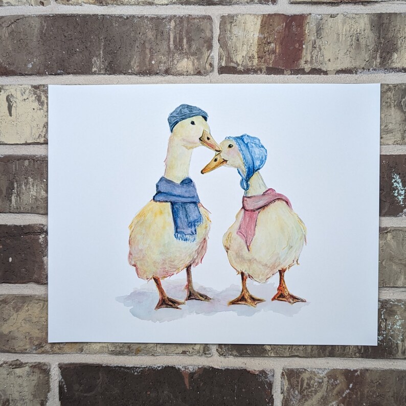K&ouml;nnte beinhalten: Zwei gelbe Enten, die blaue und rosafarbene Schals und H&uuml;te tragen. Die Enten schauen sich an und scheinen sich zu k&uuml;ssen. Der Hintergrund ist wei&szlig;.