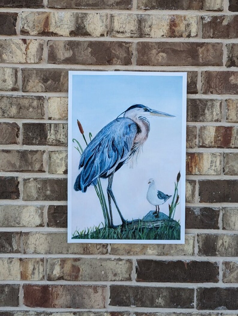 May include: A print of a blue heron standing on a rock with a smaller white bird perched on a rock nearby. The background is a light blue sky with some green reeds.