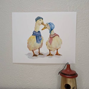 K&ouml;nnte beinhalten: Ein Aquarellbild von zwei Enten, die blaue und rosa H&uuml;te und Schals tragen. Die Enten schauen sich an und scheinen sich anzusehen.