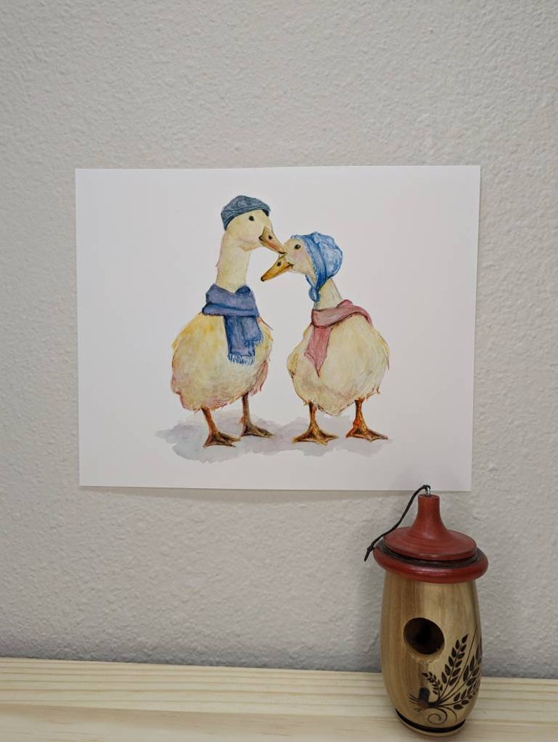 K&ouml;nnte beinhalten: Ein Aquarellbild von zwei Enten, die blaue und rosa H&uuml;te und Schals tragen. Die Enten schauen sich an und scheinen sich anzusehen.