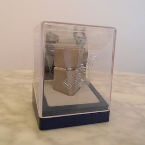 Könnte beinhalten: Eine transparente Plastikbox mit blauer Basis, die ein silbernes und goldenes Metallobjekt mit der Gravur "Versace" enthält. Die Box steht auf einer weißen Marmorfläche.