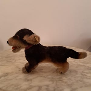 Könnte beinhalten: Ein Vintage-Stoffhundespielzeug mit einem schwarz-braunen Fell, rotem Halsband und offenem Maul. Der Spielzeughund steht auf einer Marmoroberfläche. Die Ohren des Hundes sind hellbraun und seine Augen sind dunkel.