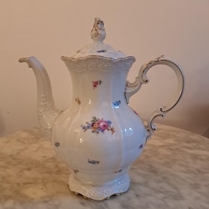 Könnte beinhalten: Weiße Porzellan-Kaffeekanne mit Deckel, Ausguss und Henkel. Die Kanne ist mit kleinen Blumensträußen in Rosa, Blau und Gelb verziert. Henkel und Rand haben goldfarbene Akzente. Die Kanne steht auf einem dekorativen Sockel.