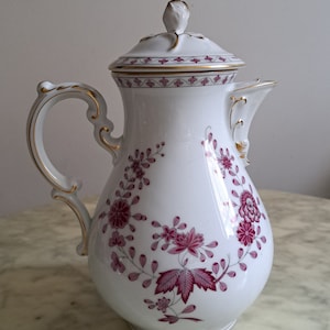 Könnte beinhalten: Weiße Porzellan-Kaffeekanne mit Deckel, verziert mit einem rosa Blumenmuster und goldenen Akzenten. Die Kanne hat einen geschwungenen Griff und einen Ausguss. Ein dekoratives Stück zum Servieren von Getränken, mit einem Fassungsvermögen von ca. 1,5 Litern.