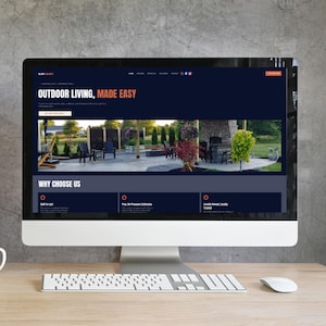 Könnte beinhalten: Ein Computermonitor zeigt eine Website mit der Überschrift Outdoor Living, Made Easy. Der Bildschirm zeigt ein Foto einer Steinterrasse mit Gartenmöbeln, einem Kamin und Landschaftsgestaltung. Eine weiße Tastatur und Maus auf einem Holztisch.
