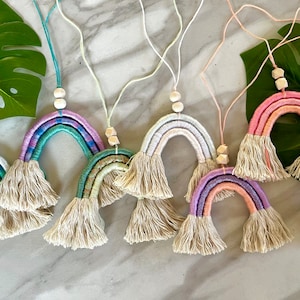 Puede incluir: Una colección de colgantes de pared hechos a mano con forma de arcoíris. Cada arcoíris presenta un arco colorido tejido con flecos de borlas y cuentas de madera. Los arcoíris son de varios colores pastel, incluyendo rosa, azul y morado.