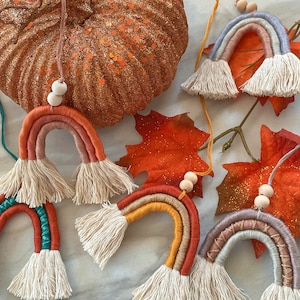 Może przedstawiać: Ręcznie robione ozdoby w kształcie tęczy z makramy w ciepłych kolorach, takich jak pomarańczowy, czerwony i żółty. Każda tęcza ma frędzel na dole i drewniane koraliki. W tle brokatowa dynia i jesienne liście.