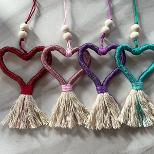 以下が含まれることがあります： 赤、ピンク、紫、ティール色の4つのハート型オーナメント。各ハートは、お揃いの糸で包まれ、下部にタッセルが付いています。木製のビーズが各ハートの上に紐で吊り下げられています。