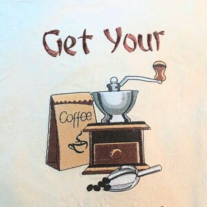May include: A brown and white kitchen towel with the text "Get Your Grind On!" embroidered on it. The towel features a brown coffee bag with the word "Coffee" printed on it, a silver coffee grinder, and a brown wooden scoop.
