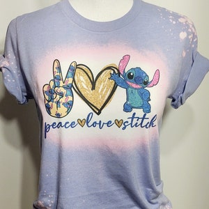 May include: A light blue t-shirt with a bleach-splattered design. The shirt features a peace sign, a heart, and a cartoon character with the text "peace love stitch".