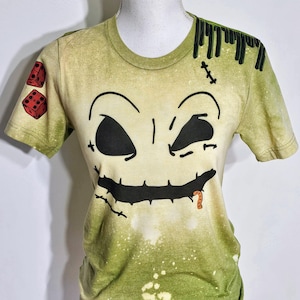 May include: A light green short-sleeve t-shirt with a bleached effect, featuring a large black stitched face graphic on the front. The right sleeve displays two red dice, while the left shoulder area shows black vertical lines and a stitch mark.