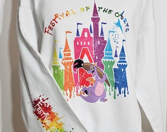 May include: White sweatshirt featuring a colourful graphic of a castle and a purple dragon holding a paintbrush. The text "Festival of the Arts" is printed in a circle around the castle. The sleeve has a rainbow paint splatter design with the text "Festival of the Arts" printed on it.