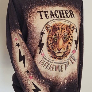 May include: Dark gray sweatshirt with a bleached design featuring a leopard's face inside a circle with the words "TEACHER DIFFERENCE MAKER". The sleeves have lightning bolts and stars.