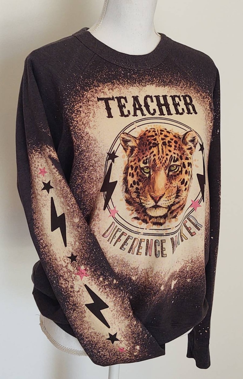 May include: Dark gray sweatshirt with a bleached design featuring a leopard's face inside a circle with the words "TEACHER DIFFERENCE MAKER". The sleeves have lightning bolts and stars.