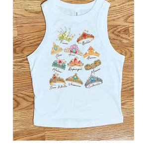 Peut inclure: D&eacute;bardeur blanc avec des illustrations color&eacute;es de diad&egrave;mes de princesses. Chaque diad&egrave;me est &eacute;tiquet&eacute; avec le nom d'une princesse, notamment Tiana, Belle, Elsa, Ariel, Aurora, Mulan, Raiponce, Moana, Blanche-Neige, Jasmine et Cendrillon.