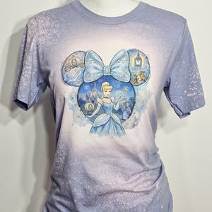 Peut inclure: T-shirt bleu clair avec un motif Cendrillon. Le motif est en forme d'oreilles de Mickey Mouse, avec Cendrillon au centre. Le t-shirt a un effet d&eacute;lav&eacute; et blanchi, avec un d&eacute;grad&eacute; de bleu clair et blanc.