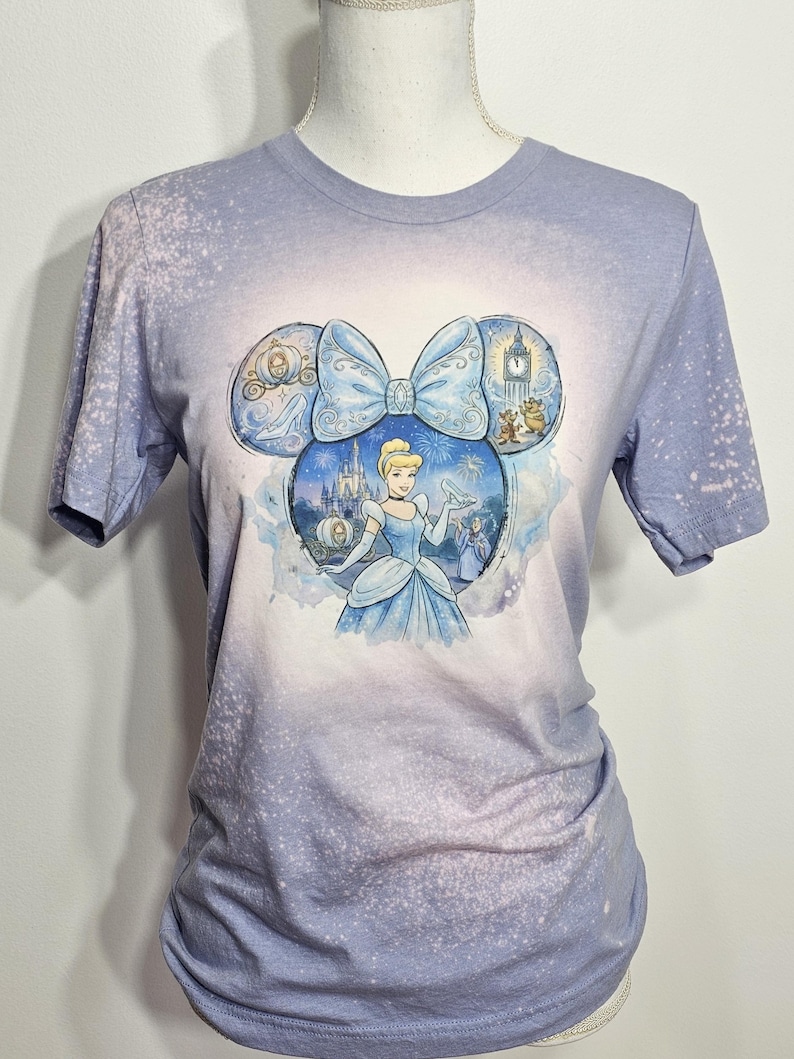 Peut inclure: T-shirt bleu clair avec un motif Cendrillon. Le motif est en forme d'oreilles de Mickey Mouse, avec Cendrillon au centre. Le t-shirt a un effet d&eacute;lav&eacute; et blanchi, avec un d&eacute;grad&eacute; de bleu clair et blanc.