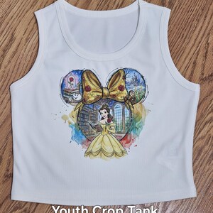 Peut inclure: D&eacute;bardeur court blanc pour jeunes avec un motif color&eacute; de Belle de La Belle et la B&ecirc;te, un n&oelig;ud jaune et des illustrations du ch&acirc;teau et de la rose. Le texte "Youth Crop Tank" est imprim&eacute; en bas.