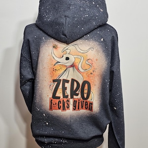 May include: A dark grey hoodie with a white splatter pattern. The back of the hoodie features a cartoon image of Zero from The Nightmare Before Christmas holding a jack-o-lantern. The text "ZERO f*cks given" is printed below the image.