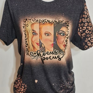Puede incluir: Una camiseta negra con efecto blanqueado y una impresi&oacute;n gr&aacute;fica de las tres hermanas Sanderson de la pel&iacute;cula Hocus Pocus. El gr&aacute;fico est&aacute; enmarcado con un borde de estampado de leopardo y el texto "Hocus Pocus" est&aacute; impreso debajo de la imagen. La camiseta tiene un dise&ntilde;o de estampado de leopardo blanqueado en las mangas.